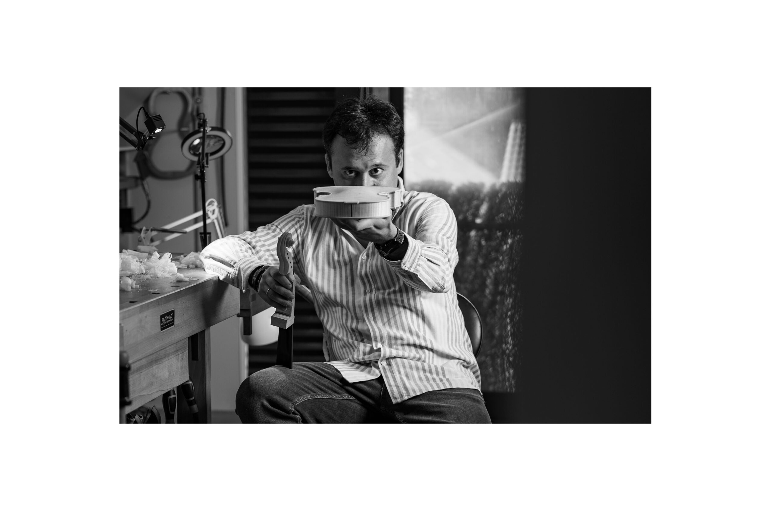 Stefan Marinescu trabajando en un violín en su taller, sujetándolo con una mano a la altura de la nariz y mirándolo concentrado en blanco y negro